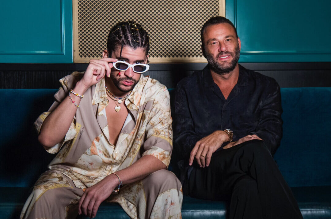 Bad Bunny and David Grutman sit side by side on a velvet bench at GEKKŌ, with Bad Bunny adjusting his signature white sunglasses and David smiling confidently, both dressed in relaxed, stylish attire.